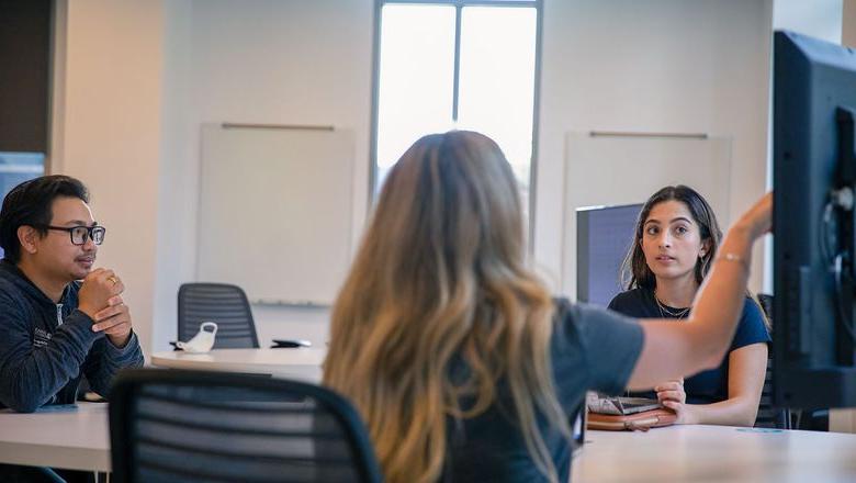 Three students engage in a discussion in front of a screen in a conference room.